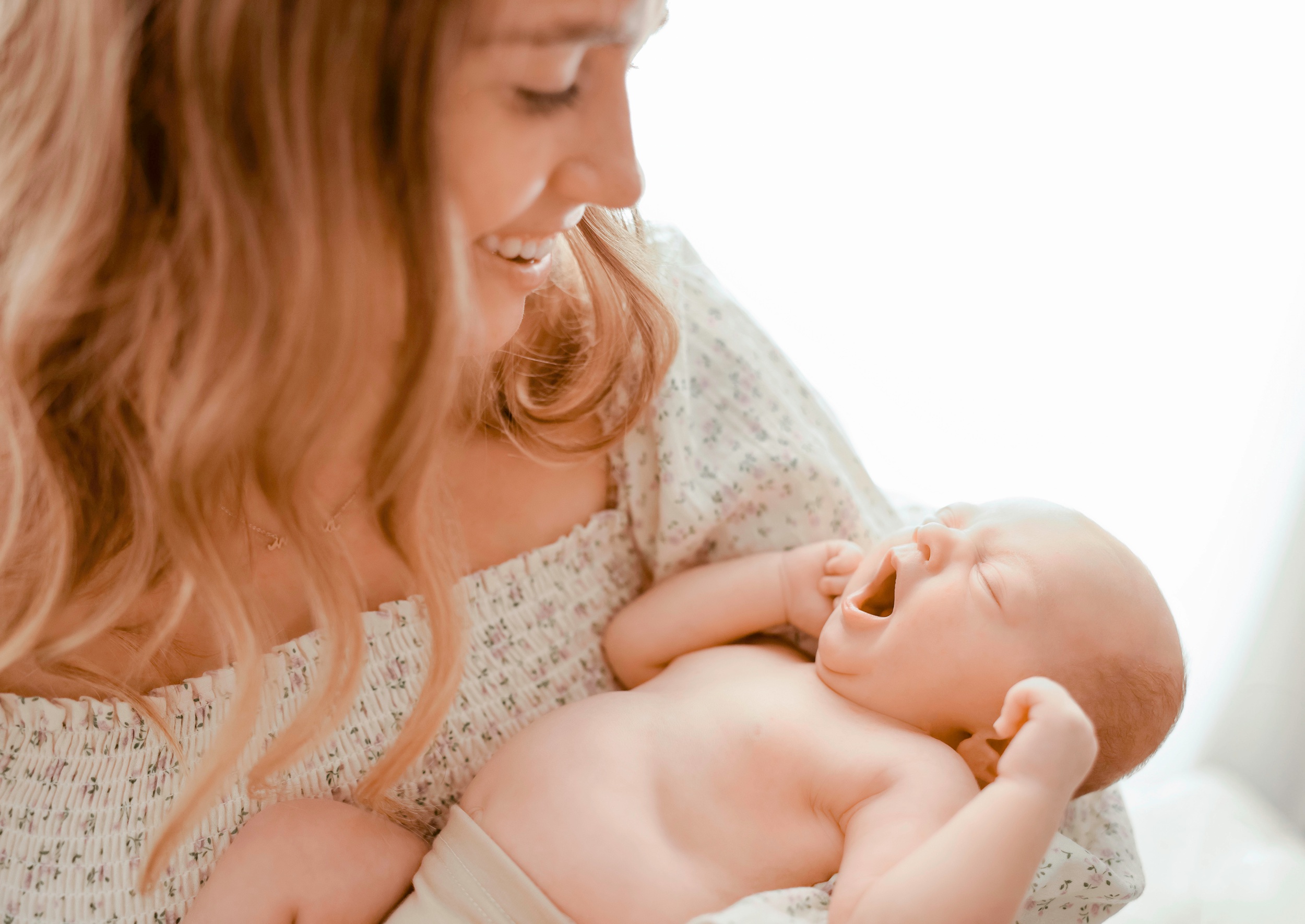 A Newborn Baby Yawns While Laying In Mom's Arms Against Her Chest In A Window After Using Maternity Clothes In Asheville