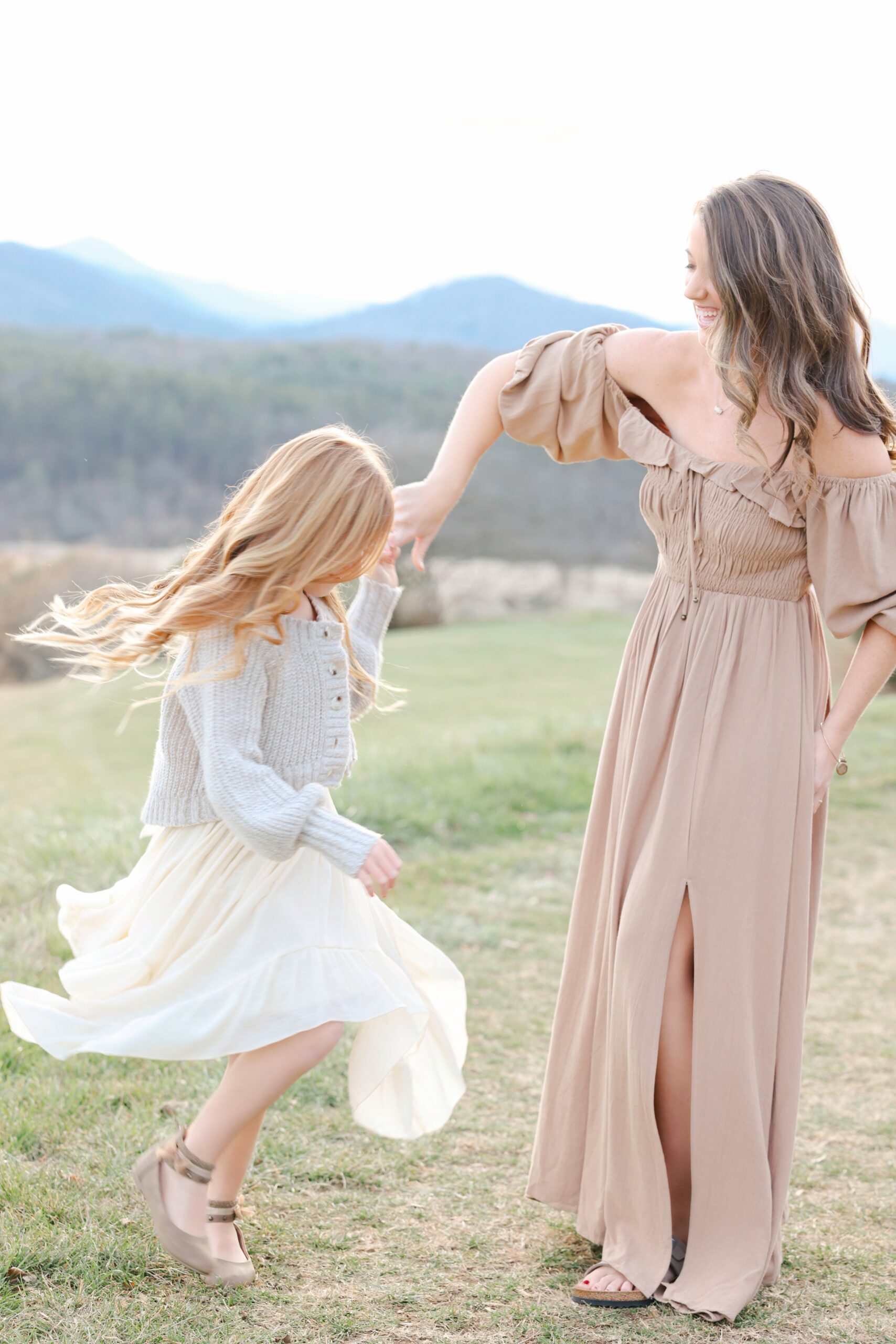 A Laughing Mom In A Brown Dress Spins Her Dancing Daughter In A Mountain Meadow