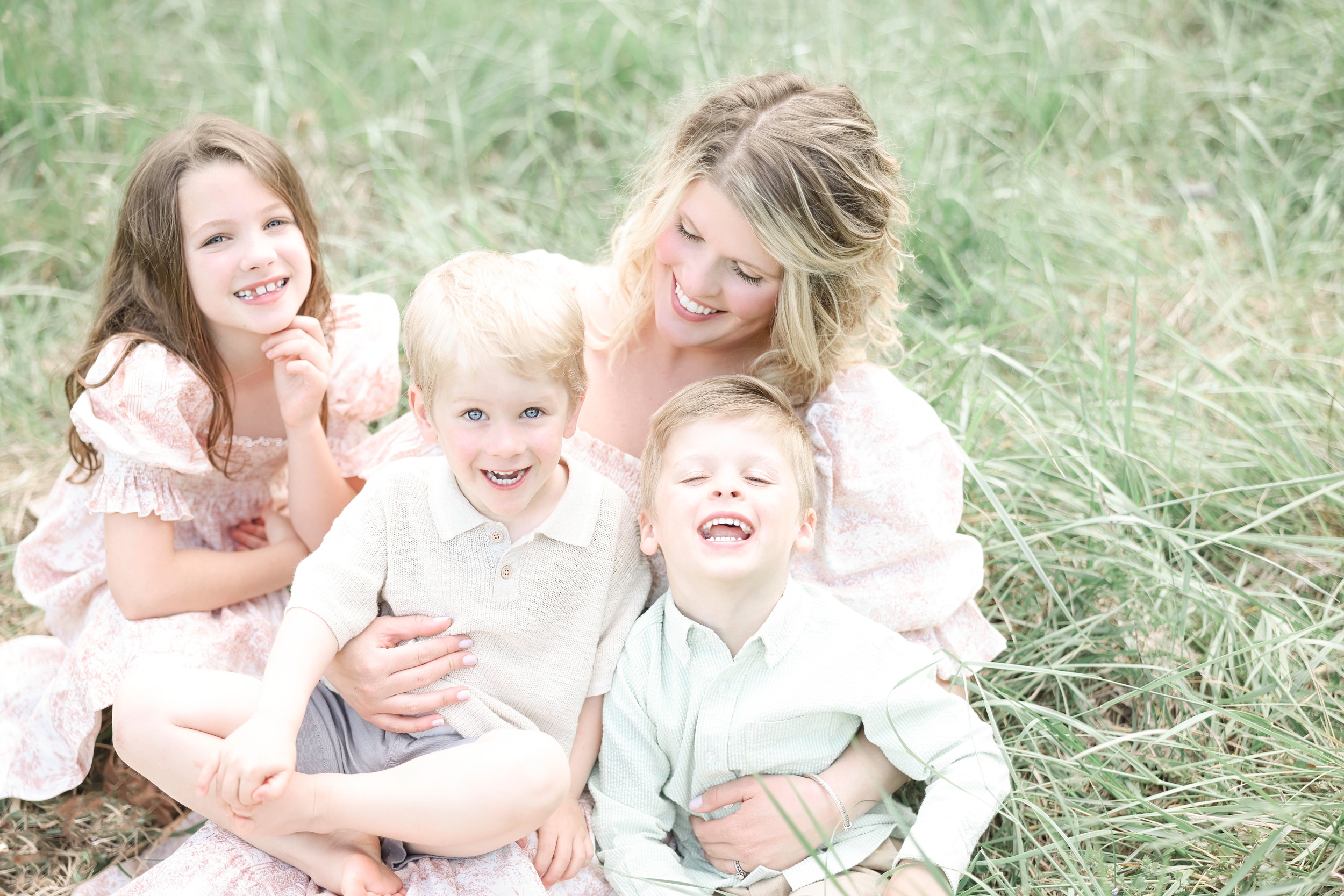 Three Laughing Toddler Siblings Sit In Mom'S Lap In A Field Of Tall Grass